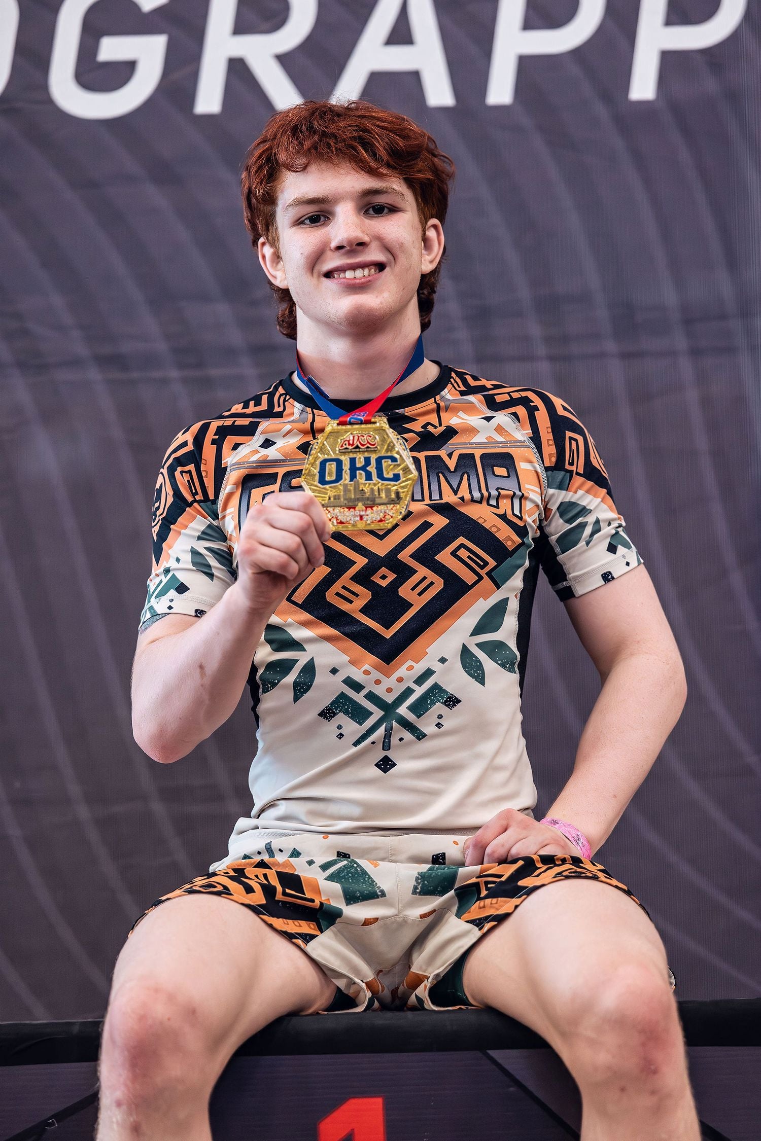 Young man wearing a tan and orange Gaidama nogi grappling set holding up his gold medal at an ADCC Open Jiu Jitsu tournament sitting on the championship podium and smiling at the camera.