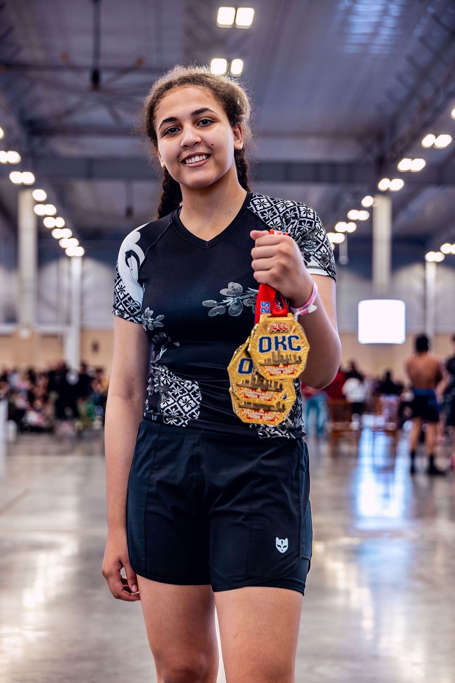 Young woman holding up 4 gold medals at an ADCC Open Jiu Jitsu competition wearing a floral black belt ranked women's rashguard and black board shorts by Gaidama with a blurry crowd of spectators in the background.