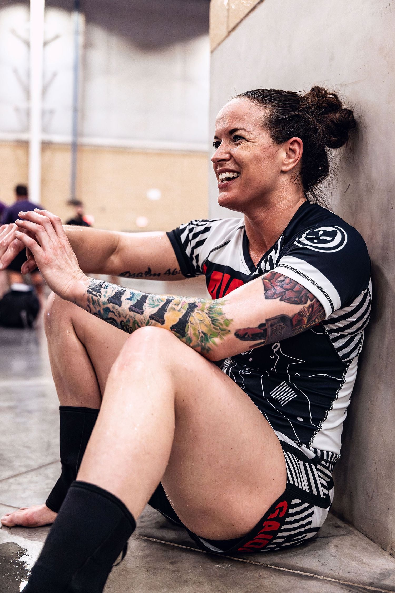 Muscular, sweaty, BJJ black belt woman sitting by a wall and smiling after competing in an ADCC Open Jiu Jitsu tournament wearing Gaidama nogi grappling apparel.