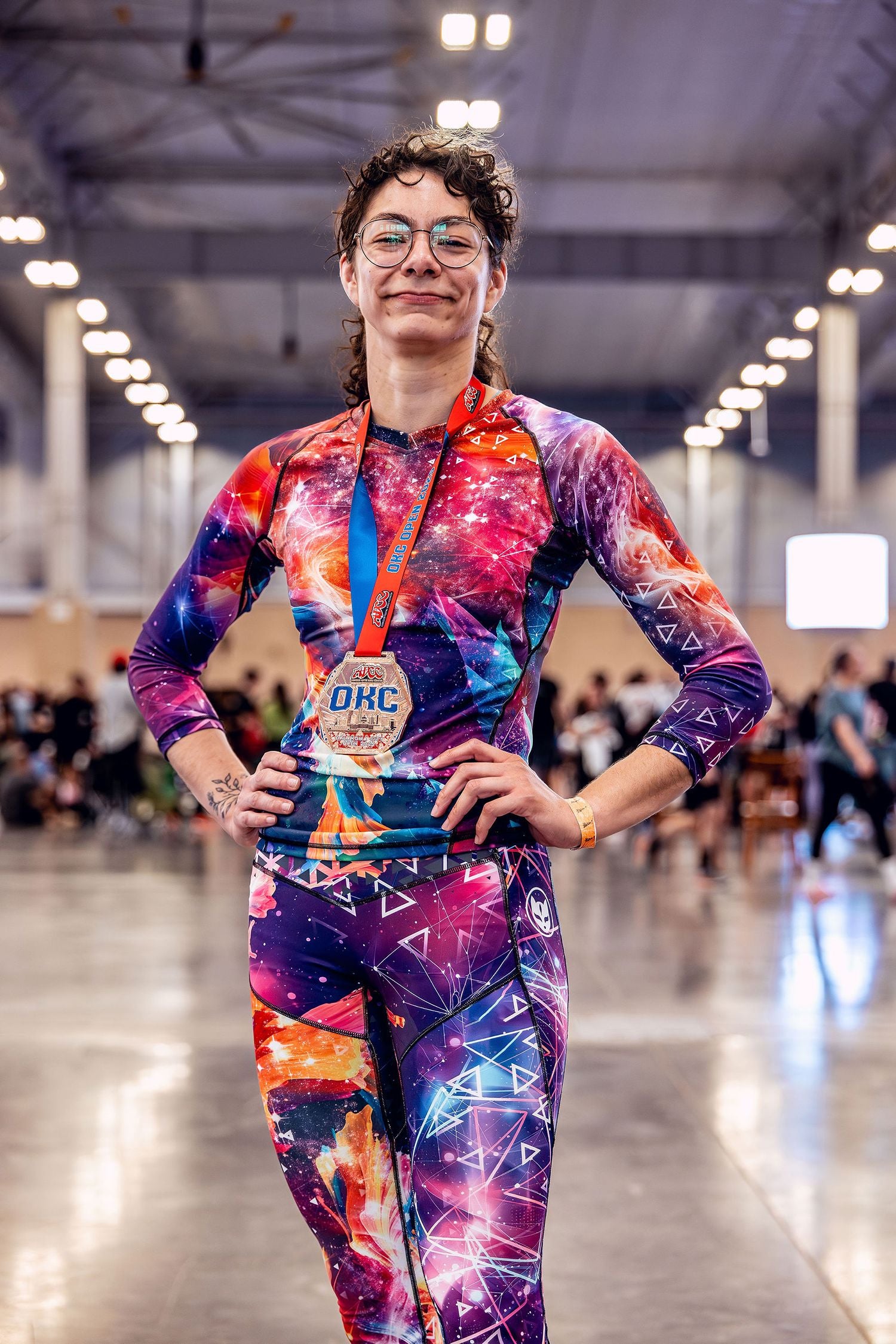 Woman standing in focus with bright lighting wearing Gaidama's pink and purple Universe Within BJJ rashguard and spats for women at an ADCC Open Jiu Jitsu tournament with a blurry crowd in the background.